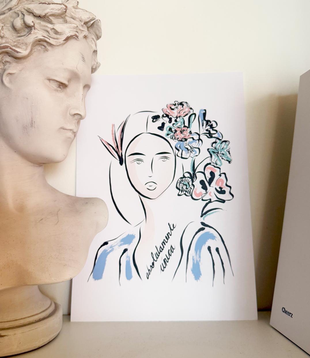 Affiche illustrée représentant une femme au regard serein, ornée de fleurs colorées dans les cheveux. Texte manuscrit "Absolumente única" sur le buste. À côté, une sculpture classique. Illustration par Leonor Roversi, à Lyon.