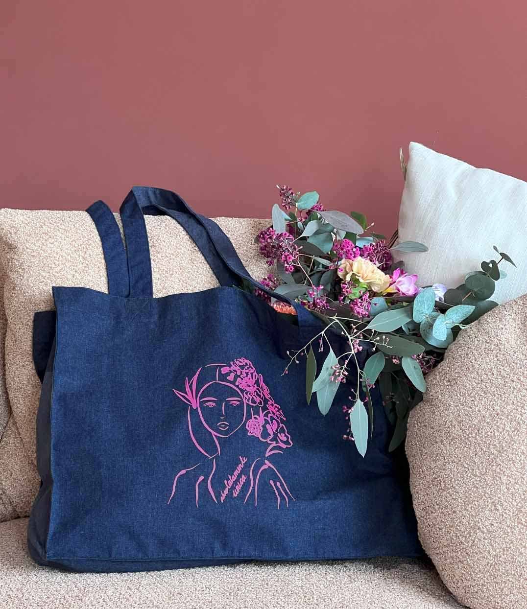 Cabas en coton bio en denim, imprimé d'une femme au regard serein entourée de fleurs dans les cheveux accompagnée de l'inscription fuchsia "Absolumente única". Modèle Unica, imprimé à Lyon par la marque éthique Leonor Roversi