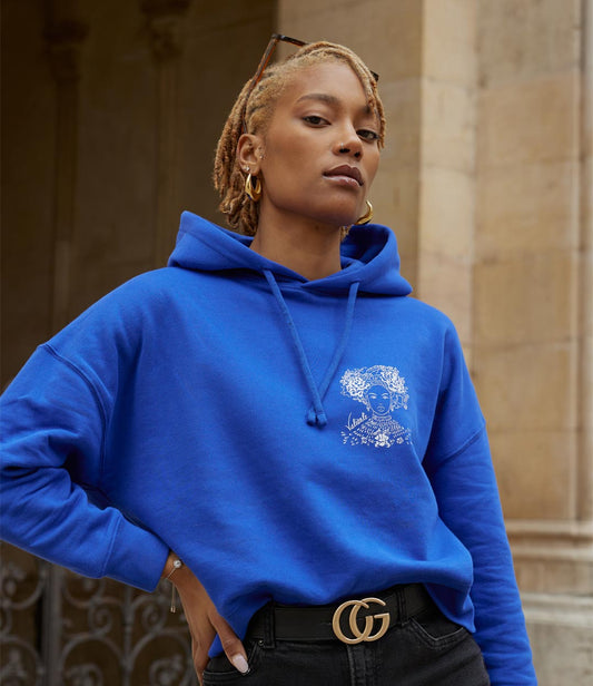 Femme portant un hoodie bleu électrique en coton biologique, imprimé blanc d'une femme forte avec des fleurs dans les cheveux représentant la première reine nigériane accompagnée de l'inscription  "Valiente" sur le côté. Modèle Valiente, conçu et imprimé à Lyon par la marque éthique Leonor Roversi.