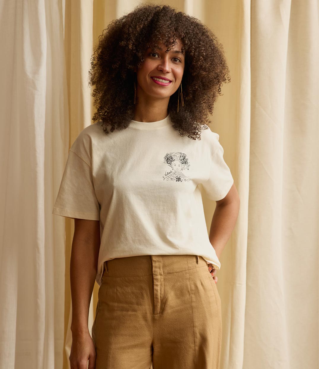 Une femme inspirante portant un t-shirt écru Valiente en coton bio de Leonor Roversi, marque lyonnaise de mode engagée et féministe, avec une illustration inspirée d’Amina de Zaria, reine nigériane symbole de puissance et d’émancipation.