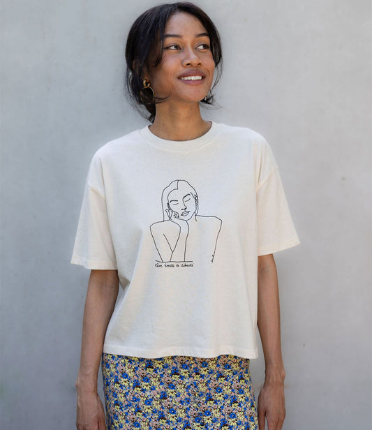Femme portant un t-shirt ample en coton bio écru. Le vêtement présente un imprimé noir minimaliste d’un visage féminin aux yeux clos, accompagné de l’inscription « rêve éveillé de liberté ». Modèle Thémis, conçu et imprimé à Lyon par la marque éthique Leonor Roversi.