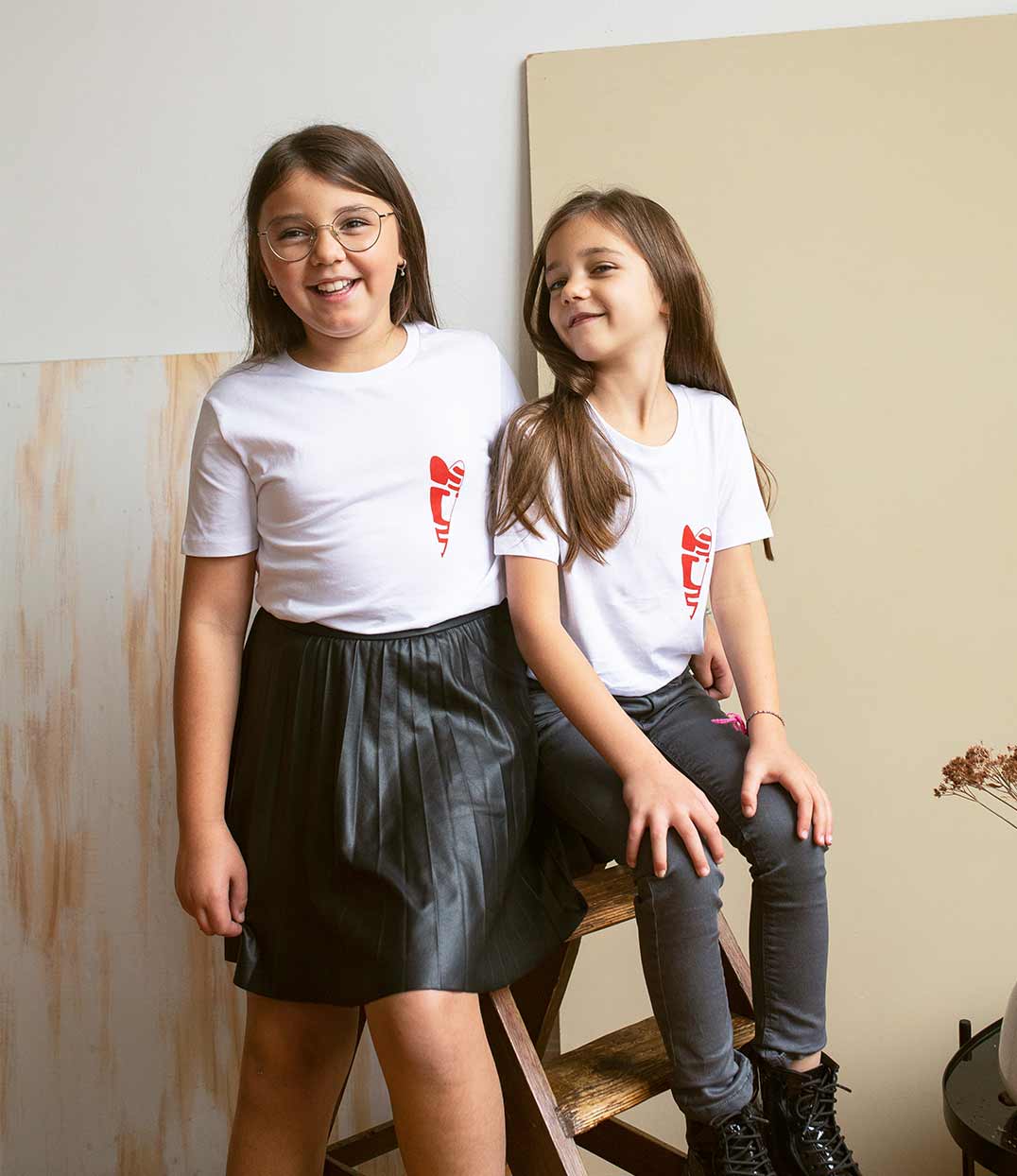 Deux enfants portent des t-shirts blancs en coton bio, ornés d’un imprimé rouge minimaliste signifiant « petit câlin » en espagnol. Une scène complice et joyeuse qui célèbre l’amour, la tendresse et l’inclusivité. T-shirts conçus dans une démarche éthique et responsable par la marque lyonnaise Leonor Roversi.