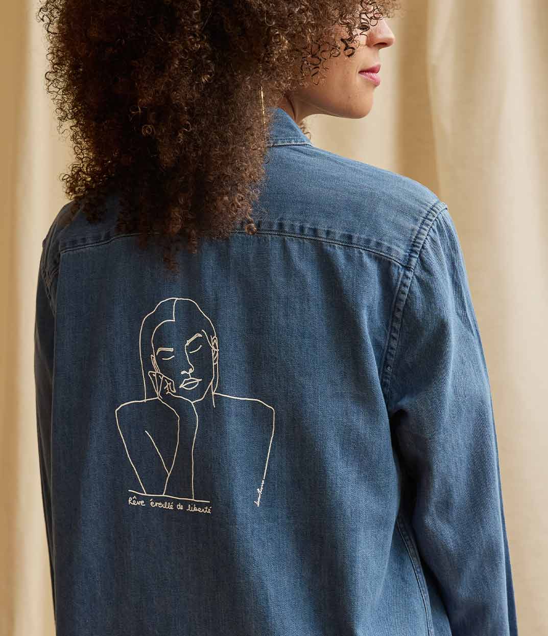 Gros plan sur une femme portant une chemise ample en coton bio denim. Le vêtement présente à l'arrière un imprimé blanc minimaliste d’un visage féminin aux yeux clos, accompagné de l’inscription « rêve éveillé de liberté ». Modèle Thémis, conçu et imprimé à Lyon par la marque éthique Leonor Roversi.