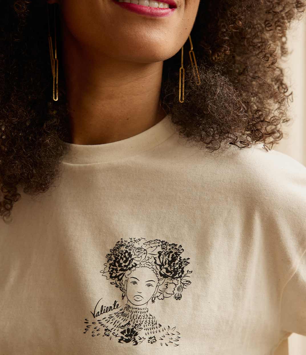 Gros plan sur une femme portant un t-shirt Valiente écru signé Leonor Roversi. Ce motif rend hommage à la reine Amina de Zaria, symbole de bravoure et d’indépendance. Une pièce engagée alliant art et mode, imaginée par une marque lyonnaise valorisant la diversité, l’artisanat local et la production durable.