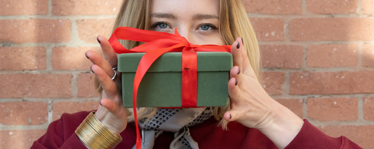 "Femme tenant une boîte cadeau verte au ruban rouge devant son visage, illustrant le thème des cadeaux de Noël éthiques. Elle porte un sweat bordeaux et des accessoires de la marque féministe Leonor Roversi."
