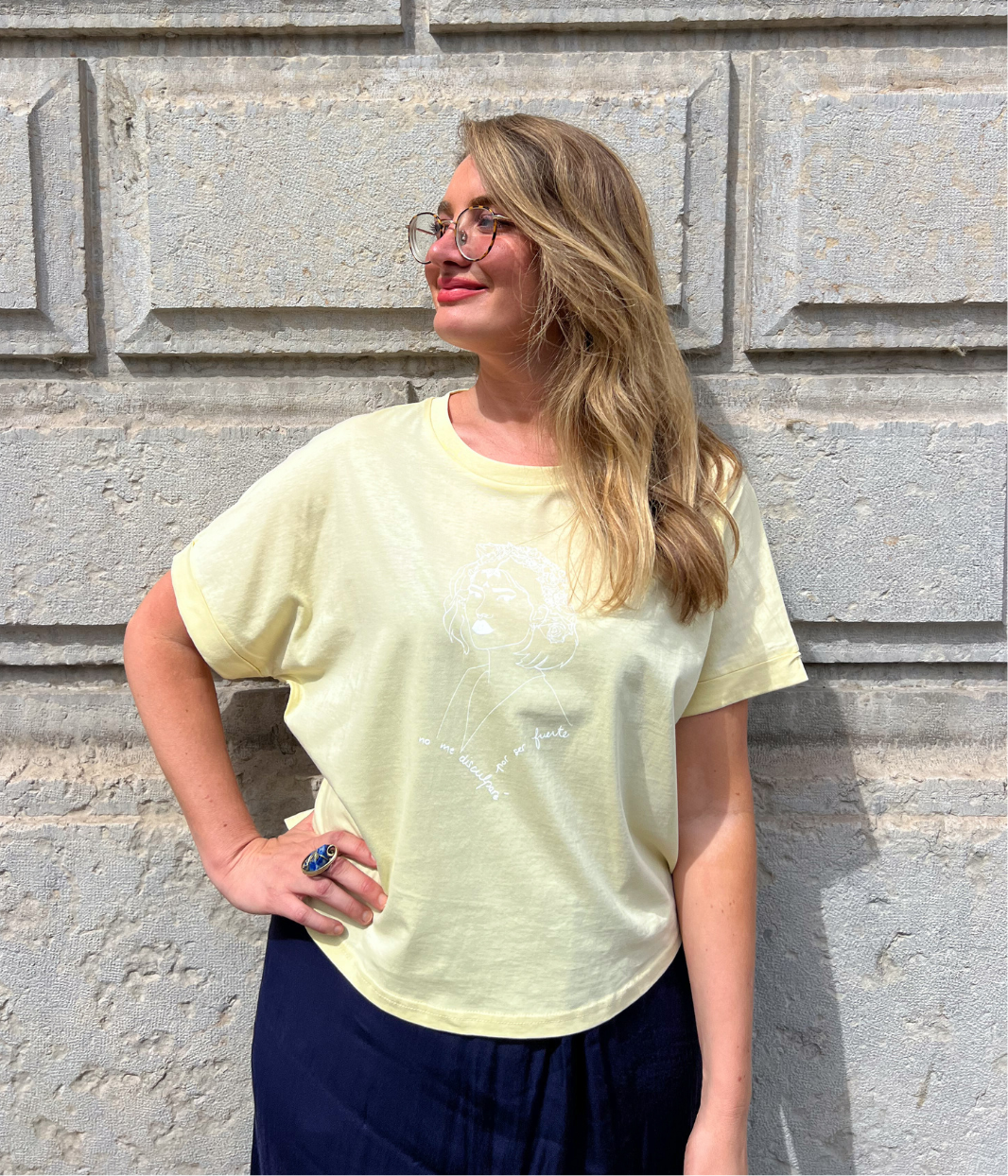 Femme blonde portant un t-shirt jaune pastel Leonor Roversi avec illustration blanche “no me disculparé por ser fuerte”, posant au soleil devant un mur en pierre.