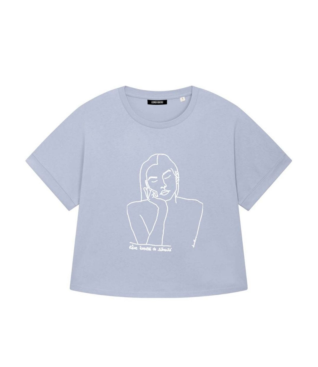 Mock-up d'un t-shirt ample pour femme en coton bio bleu serein. Le vêtement présente un à l'avant, un imprimé blanc minimaliste d’un visage féminin aux yeux clos, accompagné de l’inscription « rêve éveillé de liberté ». Modèle Thémis, conçu et imprimé à Lyon par la marque éthique Leonor Roversi.