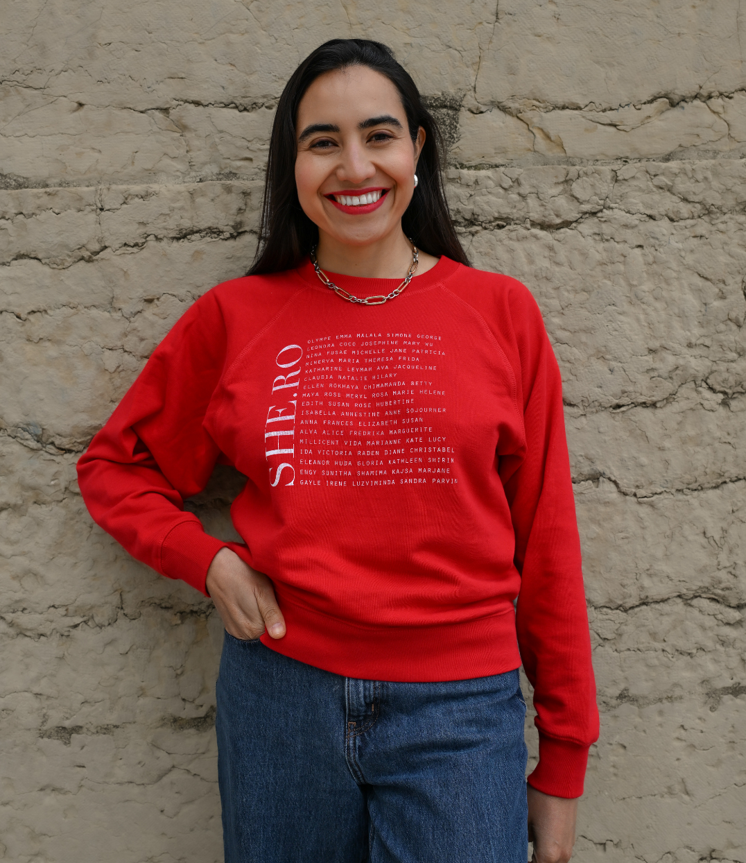 Femmes qui porte un sweat Rouge shero de chez Leonor Roversi. SHE.RO comme HERO mais pour les femmes, héroïnes. Elle est devant un mur en pierre.