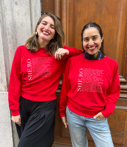 Il y a deux femmes qui portent un Sweat Shero rouge de la marque Leonor Roversi. SHE.RO comme HERO mais pour les femmes, héroïnes. Elles sont debout devant une grande porte en bois.