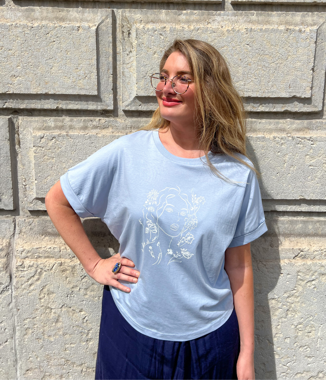 Femme aux cheveux bouclés portant un t-shirt bleu clair Leonor Roversi avec illustration blanche “Rêve éveillé de liberté”, dans une ambiance lumineuse en extérieur.
