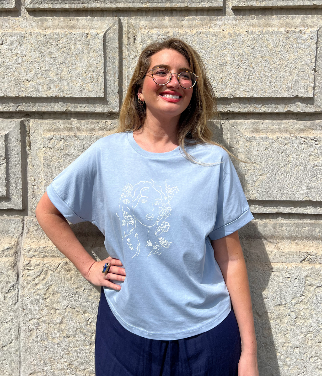 Femme blonde portant un t-shirt bleu clair Leonor Roversi avec illustration florale blanche “À l’aube de soi”, posant devant un mur en pierre.