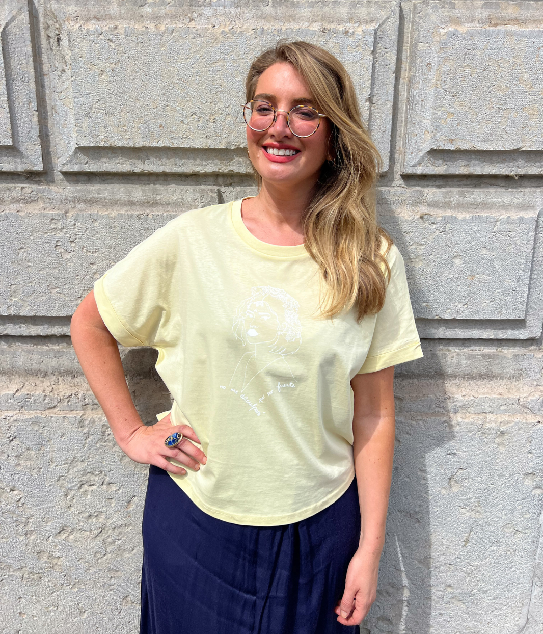 Femme souriante portant un t-shirt jaune pastel Leonor Roversi avec illustration féminine blanche, associée à un pantalon bleu marine.