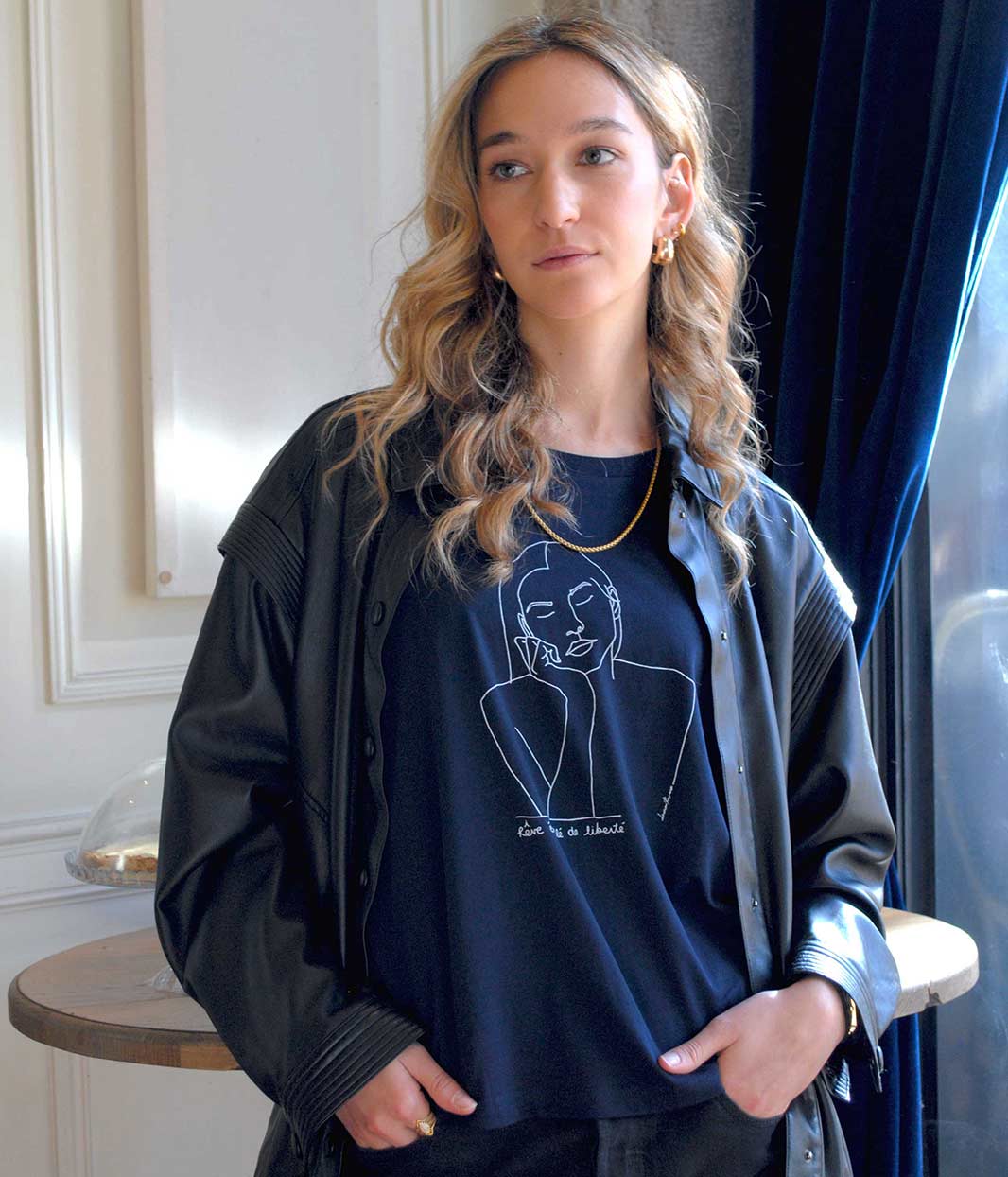Femme portant le t-shirt Thémis Leonor Roversi bleu marine en coton bio certifié GOTS et FairWear, sous une veste noire, dans un intérieur lumineux. Illustration minimaliste blanche de Thémis, symbole de justice et de bienveillance, sérigraphie éco-responsable réalisée à Lyon.