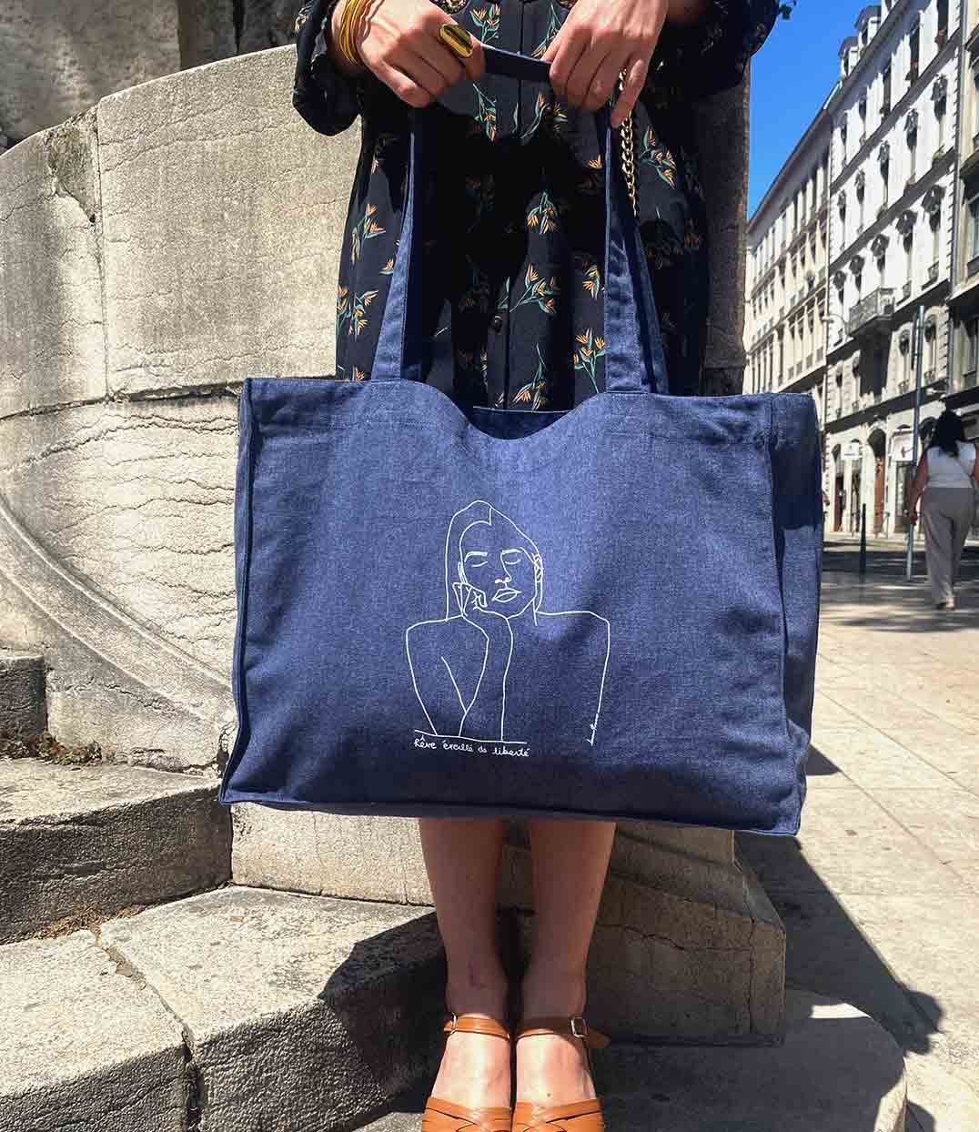 Femme portant un t-shirt ample en coton bio écru. Le vêtement présente un imprimé noir minimaliste d’un visage féminin aux yeux clos, accompagné de l’inscription « rêve éveillé de liberté ». Modèle Thémis, conçu et imprimé à Lyon par la marque éthique Leonor Roversi.