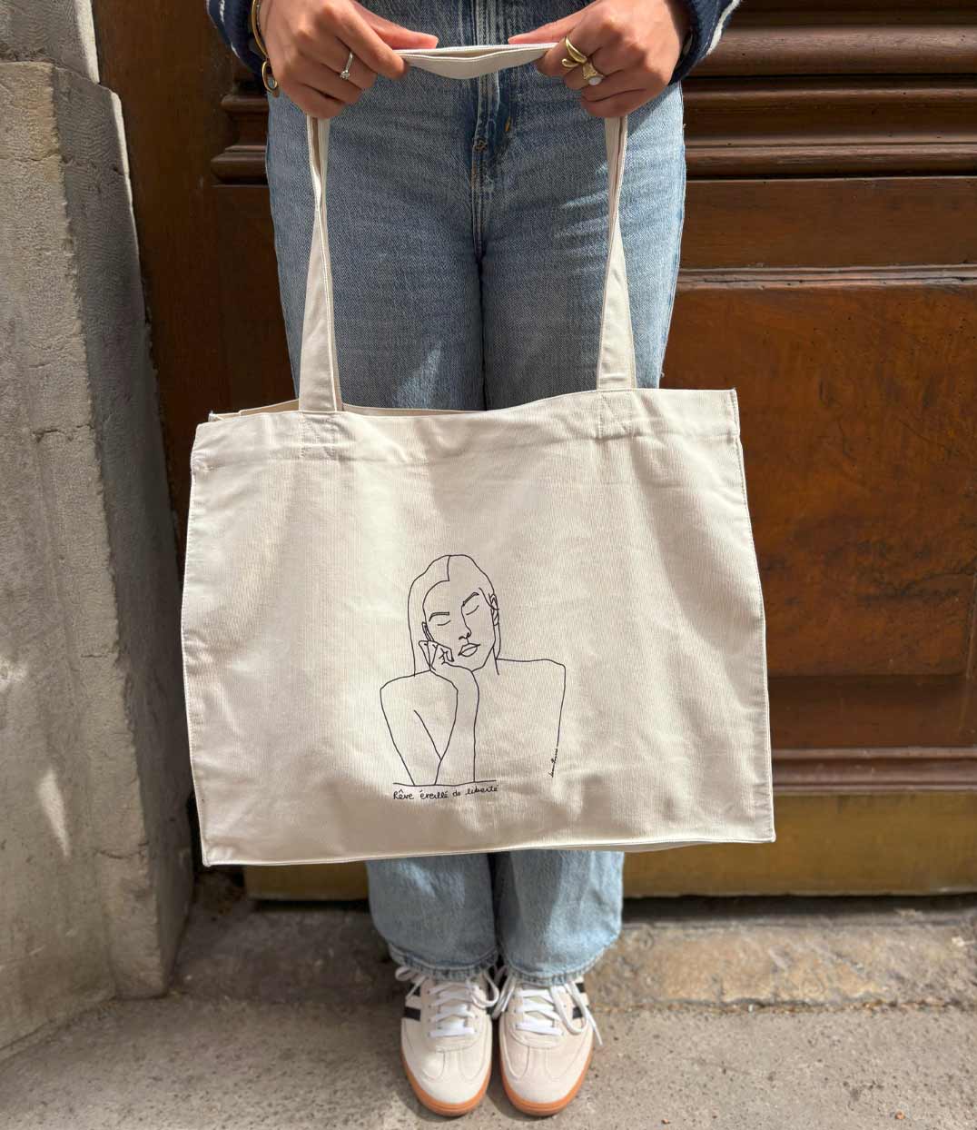 Cabas en coton bio écru, imprimé noir d'un dessin minimaliste représentant une femme sereine aux yeux fermés, accompagné de la phrase manuscrite "Rêve éveillé de liberté". Modèle Thémis, imprimé à Lyon par la marque éthique Leonor Roversi.