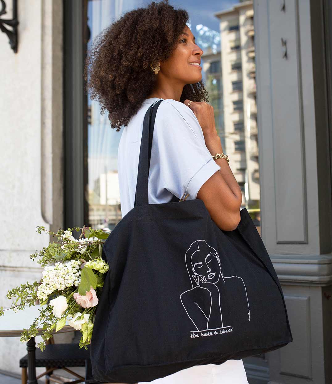 Femme portant un cabas en coton bio noir. Le vêtement présente un imprimé blanc minimaliste d’un visage féminin aux yeux clos, accompagné de l’inscription « rêve éveillé de liberté ». Modèle Thémis, conçu et imprimé à Lyon par la marque éthique Leonor Roversi.