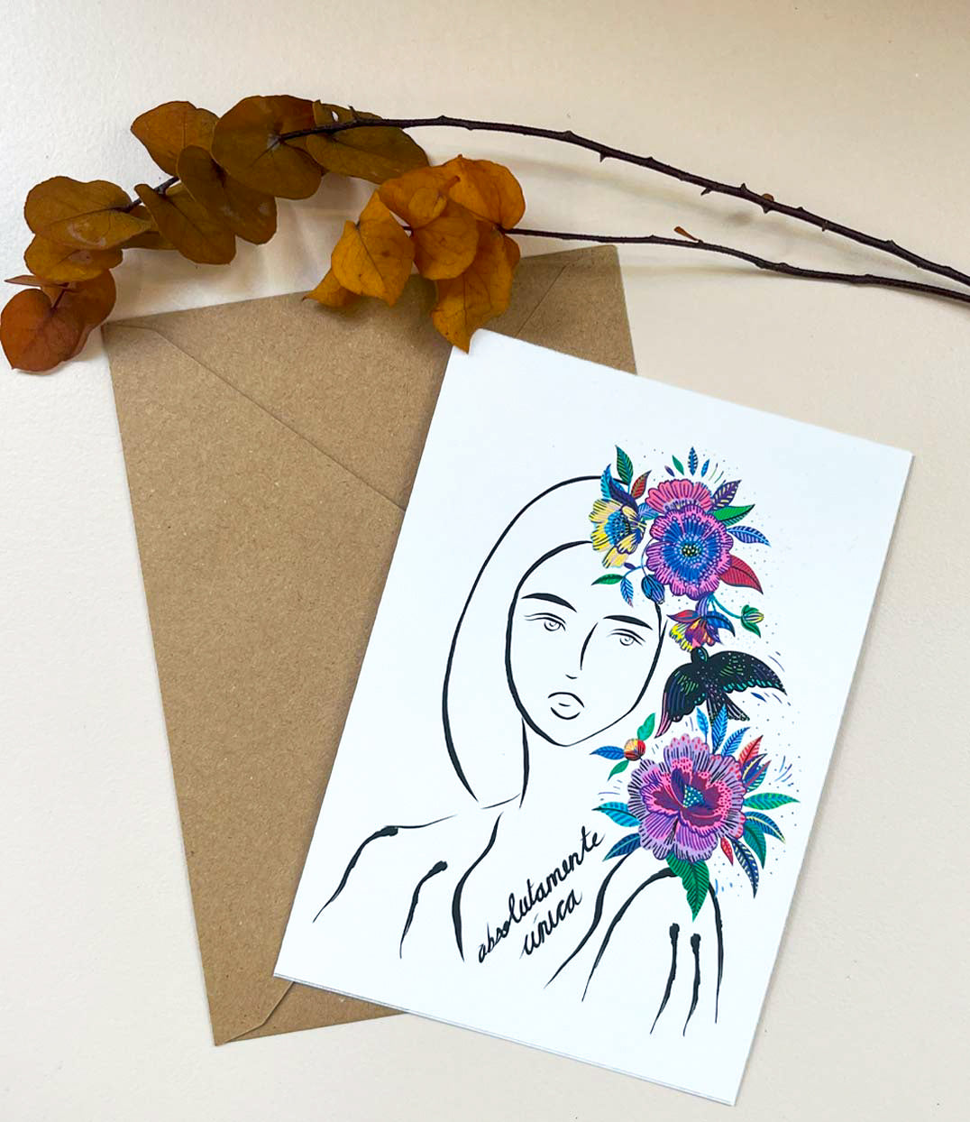 Carte postale illustrée d'une femme au regard serein entourée de fleurs colorées dans les cheveux accompagnée de l'inscription "Absolumente única" posée sur une enveloppe kraft accompagnée d'une branche. Modèle Unica en collaboration avec La Sigalerie, conçue et imprimée à Lyon par la marque éthique Leonor Roversi.