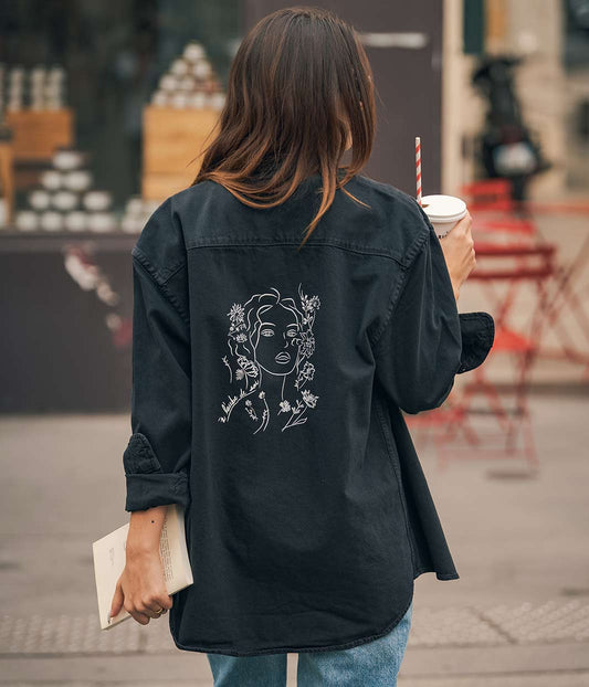 Mannequin femme de dos qui porte une chemise noire/grise avec un dessin sur le dos, Leonor Roversi. Avec un livre à la main et une boisson dans l’autre