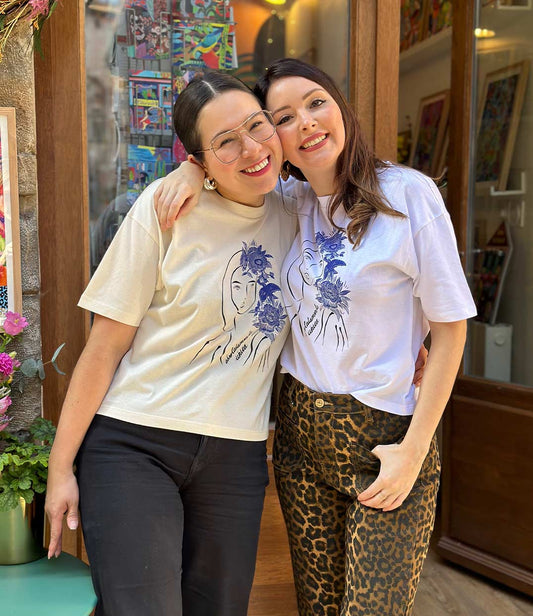 Deux femmes souriantes portant le t-shirt écru et blanc de la collection en collaboration avec La Sigalerie du dessin Unica orné de fleurs et d'animaux bleus de la marque Leonor Roversi