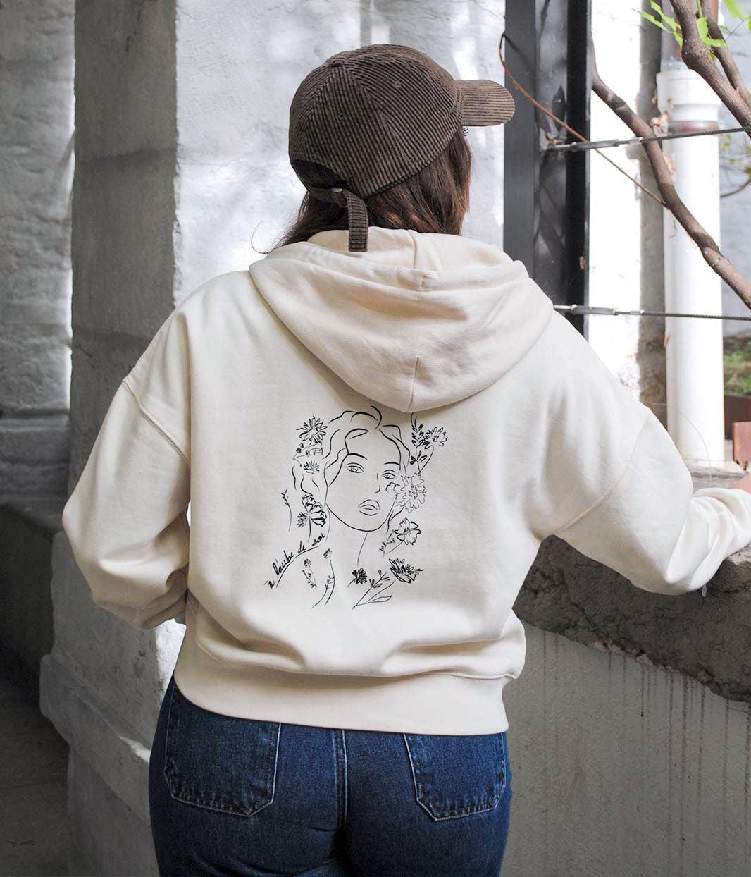 Femme photographiée de dos portant un hoodie oversize écru en coton biologique, avec imprimé noir au dos représentant un visage féminin entouré de fleurs, symbole de renouveau et de transformation. Sérigraphie artisanale éco-responsable réalisée à Lyon par la marque éthique Leonor Roversi.v