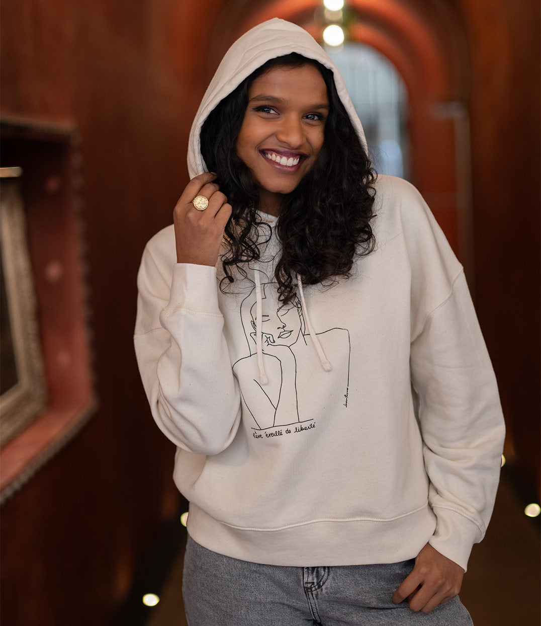 Femme portant un hoodie écru à capuche en coton bio, modèle Thémis de la marque française éco-responsable Leonor Roversi. La capuche est relevée, mettant en valeur l’imprimé minimaliste d’un visage féminin symbolisant la justice et la bienveillance. Sérigraphie artisanale réalisée à Lyon, coupe ample et moderne, sweat éthique conçu à partir de coton biologique certifié GOTS et de polyester recyclé.
