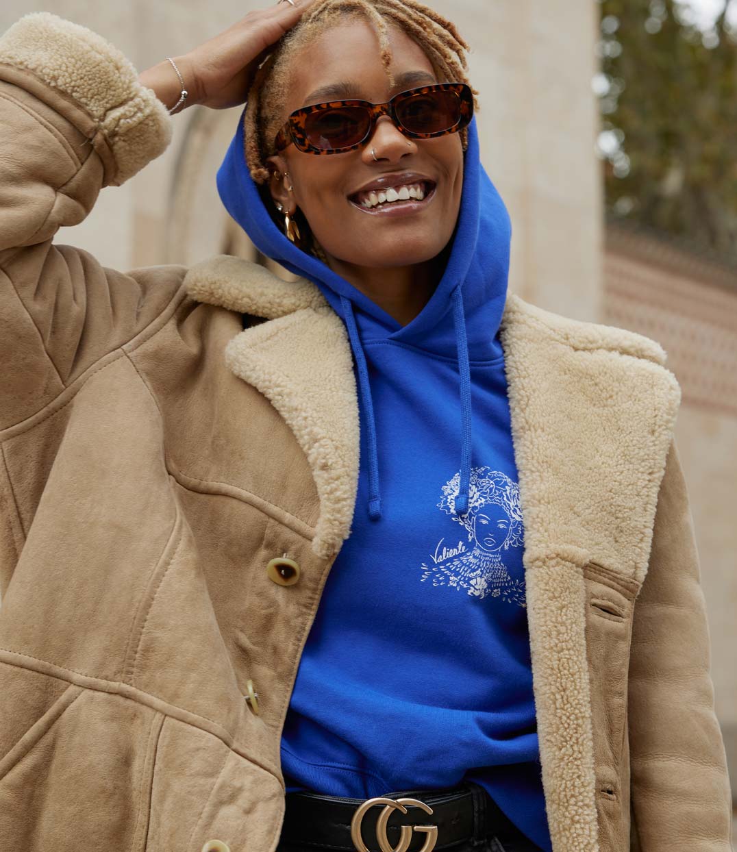 Femme portant un hoodie bleu électrique en coton biologique, imprimé blanc d'une femme forte avec des fleurs dans les cheveux représentant la première reine nigériane accompagnée de l'inscription "Valiente" sur le côté avec une veste en daim. Modèle Valiente conçu et imprimé à Lyon par la marque éthique Leonor Roversi.
