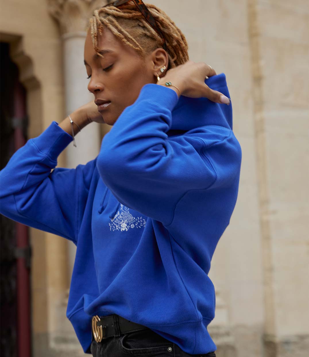 Femme portant un hoodie bleu électrique en coton biologique, imprimé blanc d'une femme forte avec des fleurs dans les cheveux représentant la première reine nigériane accompagnée de l'inscription "Valiente" sur le côté. Modèle Valiente conçu et imprimé à Lyon par la marque éthique Leonor Roversi.