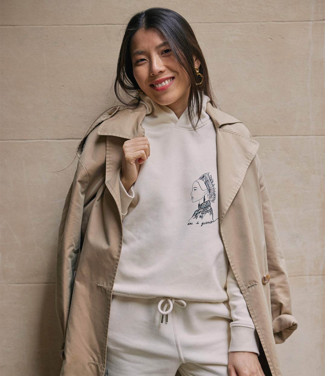 Femme souriante portant un hoodie en coton bio écru. Le vêtement présente un imprimé blanc représentant une guerrière japonaise, accompagnée de l’inscription « âme d'une guerrière ». Modèle Benten, conçu et imprimé à Lyon par la marque éthique Leonor Roversi.