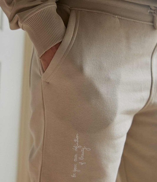 Pantalon de jogging femme en coton bio sable de Leonor Roversi, en coton biologique, avec l’inscription brodée « Be your own definition of beauty ». Pièce douce et confortable, pensée pour une mode responsable qui célèbre l’amour de soi et l’authenticité.