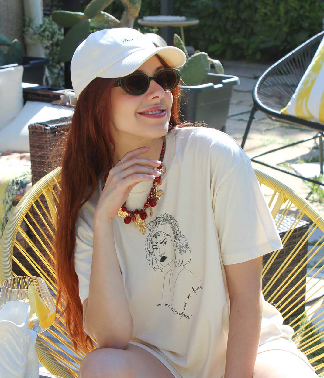 Femme assise en extérieur portant des lunettes noires et un t-shirt jaune pastel avec illustration blanche d’un visage entouré de fleurs.