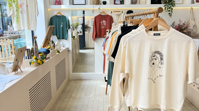 Intérieur de la boutique lyonnaise et éthique Leonor Roversi. Il y a des portants avec des t-shirts, un t-shirt vert aloe et un sweat terracotta suspendu au mur avec un miroir murale.