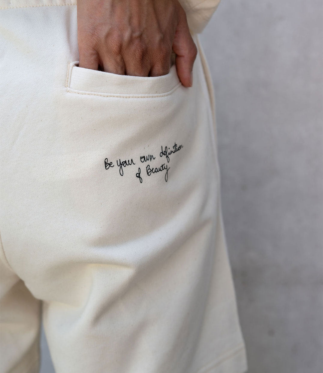 Gros plan sur l’arrière d’un short femme en coton bio écru, avec une inscription manuscrite noire : “Be your own definition of beauty”. Modèle Sekhmet, imprimé à Lyon par la marque éthique Leonor Roversi.