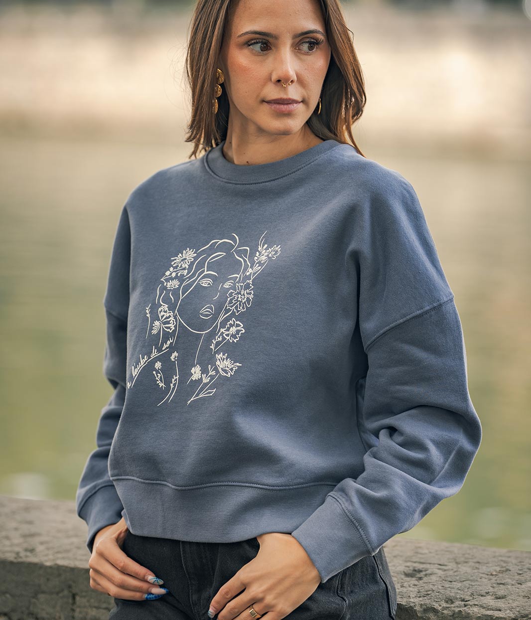Mannequin femme, les mains dans les poches, portant un sweat-shirt coton bio gris bleu, avec un dessin Aurora Leonor Roversi