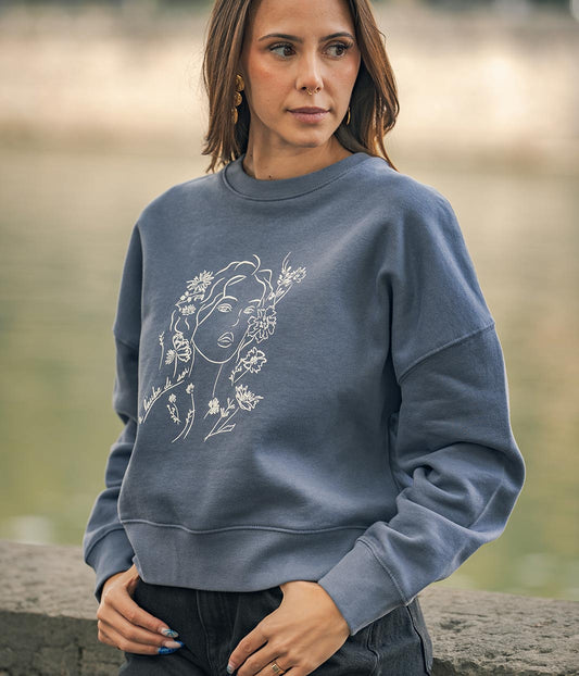 Mannequin femme, les mains dans les poches, portant un sweat-shirt coton bio gris bleu, avec un dessin Aurora Leonor Roversi