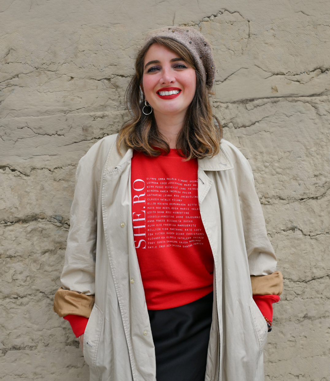 Femmes qui porte un sweat Rouge shero de chez Leonor Roversi. SHE.RO comme HERO mais pour les femmes, héroïnes. Elle est devant un mur en pierre.