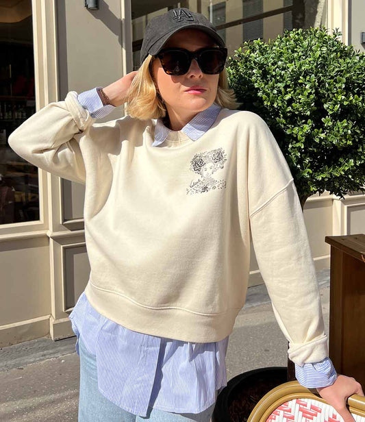 Femme portant un sweatshirt écru en coton biologique, imprimé noir d'une femme forte avec des fleurs dans les cheveux représentant la première reine nigériane accompagnée de l'inscription  "Valiente" sur le côté. Modèle Valiente conçu et imprimé à Lyon par la marque éthique Leonor Roversi.