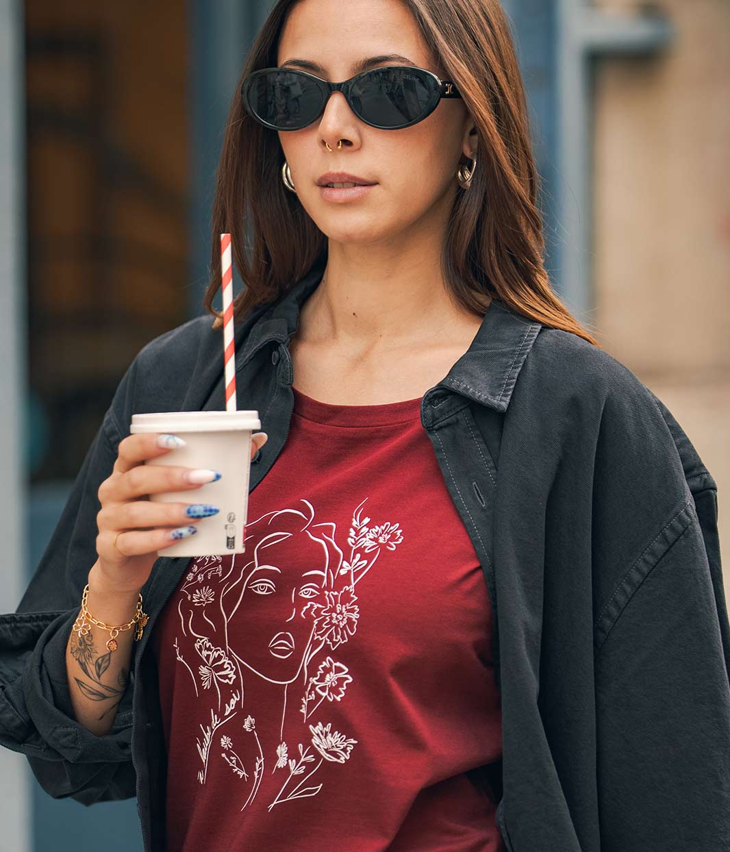 Mannequin femme, porte une chemise noire en denim et en dessous un t-shirt en coton bio, rouge, bordeaux avec un dessin dessus Leonor Roversi. Avec ses lunettes de soleil et une boisson à la main 