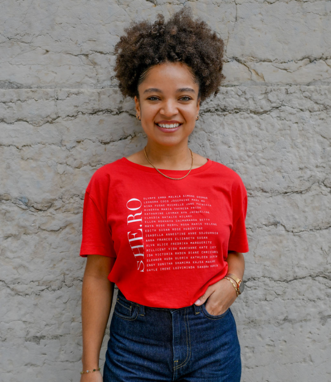 Femmes qui porte un t-shirt Rouge shero de chez Leonor Roversi. SHE.RO comme HERO mais pour les femmes, héroïnes. Elle est devant un mur en pierre.