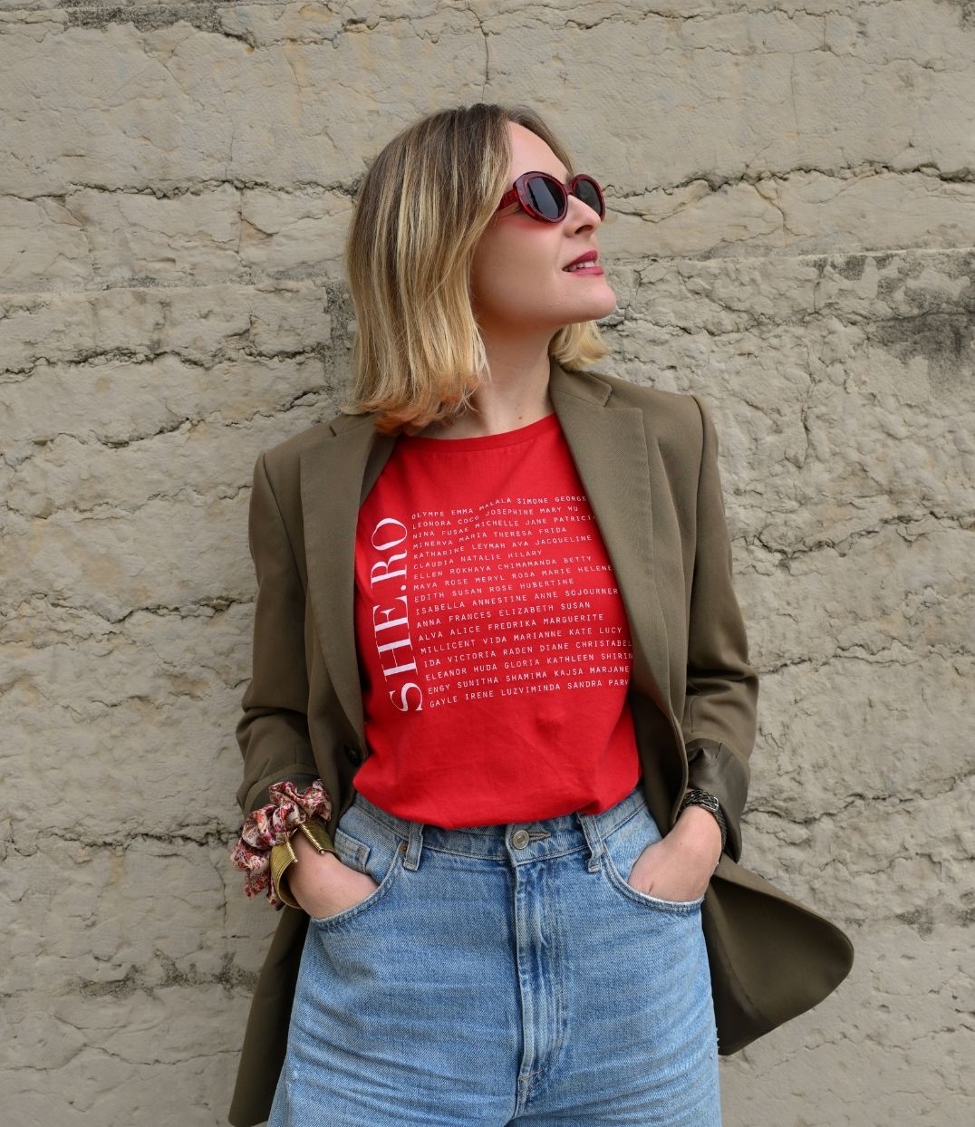 Femmes qui porte un t-shirt Rouge shero de chez Leonor Roversi. SHE.RO comme HERO mais pour les femmes, héroïnes. Elle est devant un mur en pierre.