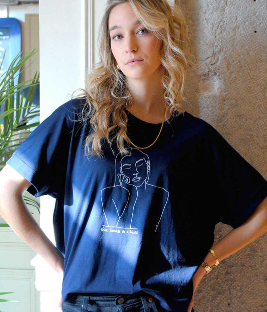 Femme portant le t-shirt Thémis Leonor Roversi bleu marine en coton bio, debout près d’un mur en pierre, mains sur les hanches. Sérigraphie blanche représentant Thémis, allégorie de la justice et de l’impartialité, impression 100 % lyonnaise, éthique et réalisée avec amour.