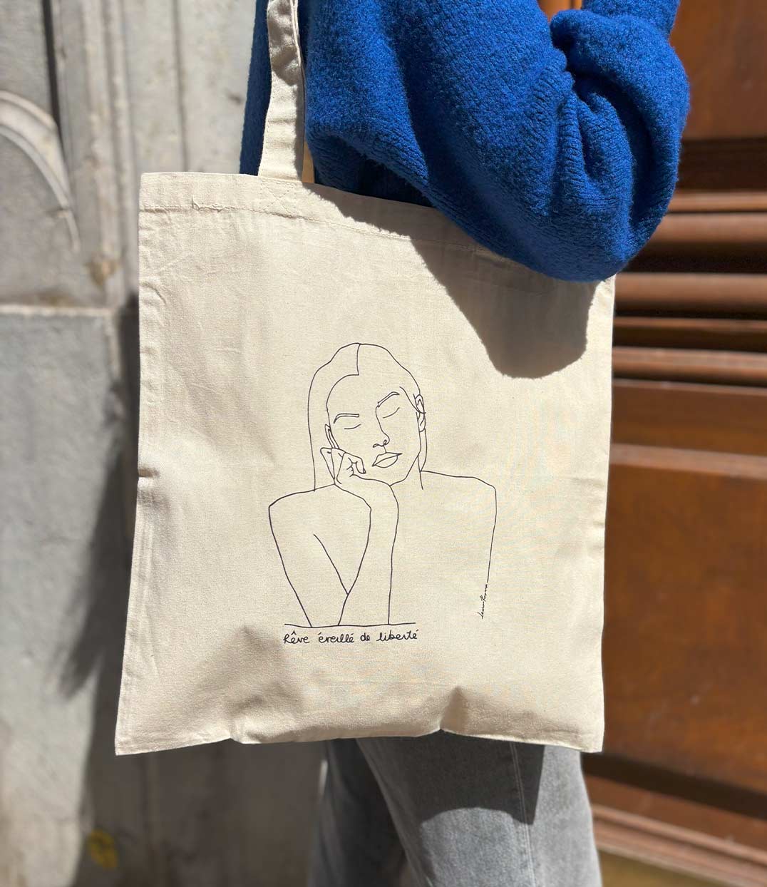 Tote Bag en coton bio écru, imprimé noir d'un dessin minimaliste représentant une femme sereine aux yeux fermés, accompagné de la phrase manuscrite "Rêve éveillé de liberté". Modèle Thémis, imprimé à Lyon par la marque éthique Leonor Roversi.