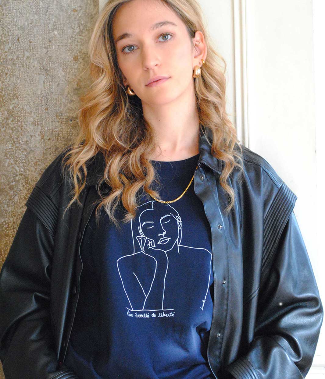 Femme portant le t-shirt Thémis Leonor Roversi bleu marine, col rond et coupe courte et droite, devant un mur clair. Dessin linéaire blanc représentant Thémis, figure mythologique de la justice, sérigraphie locale et éthique réalisée à Lyon.