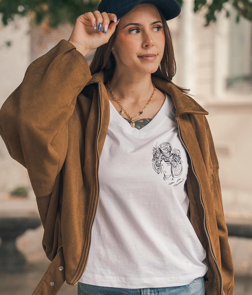Mannequin femme, tenant sa casquette bleu Leonor Roversi. Avec une veste en marron et un t-shirt col V en coton bio, blanc, Benten Leonor Roversi.
