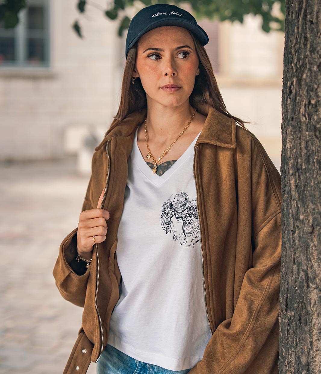 Mannequin femme, portant une casquette bleu Leonor Roversi. Avec une veste en marron et un t-shirt col V en coton bio, blanc, Benten Leonor Roversi.