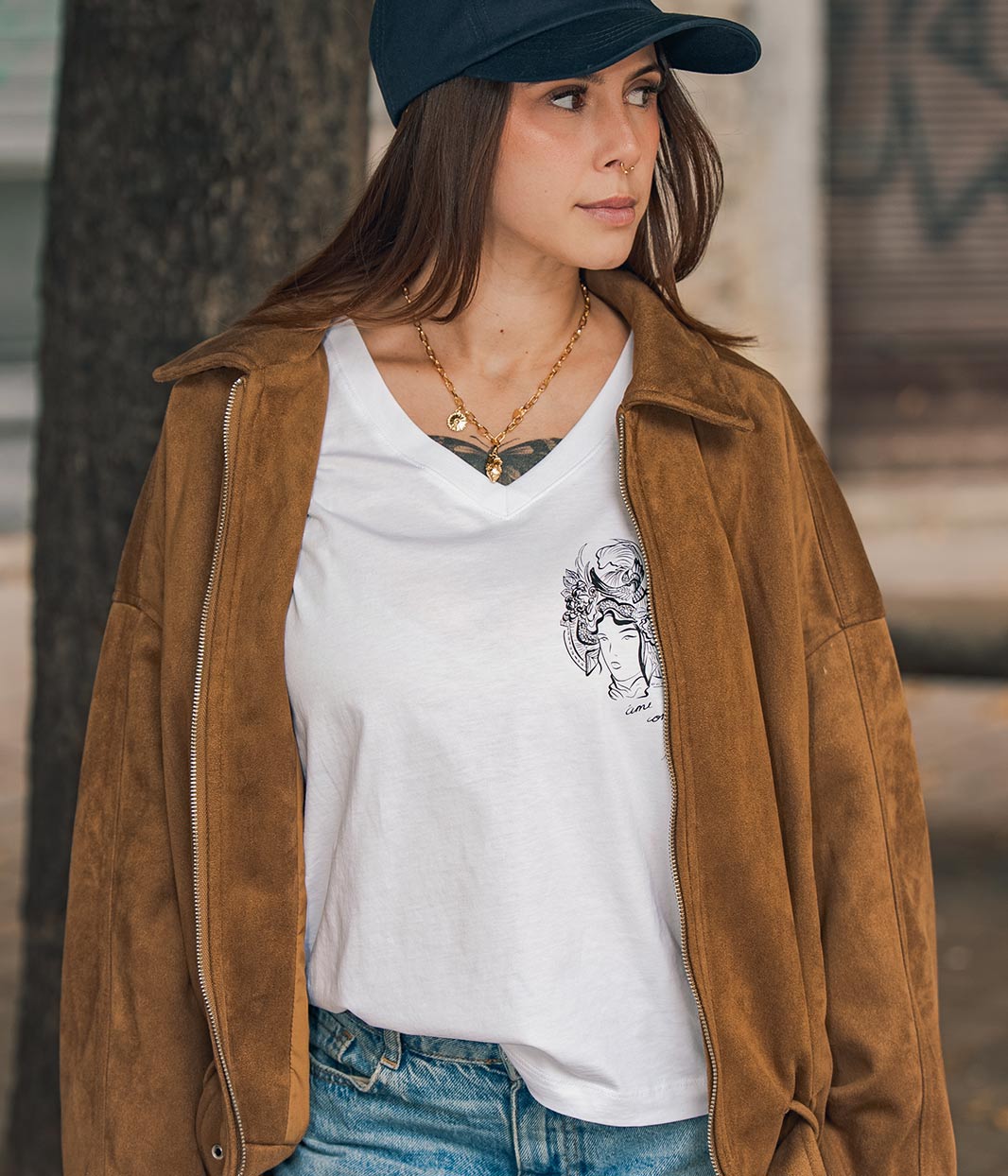 Mannequin femme, portant une casquette bleu Leonor Roversi. Avec une veste en marron et un t-shirt col V en coton bio, blanc, Benten Leonor Roversi.
