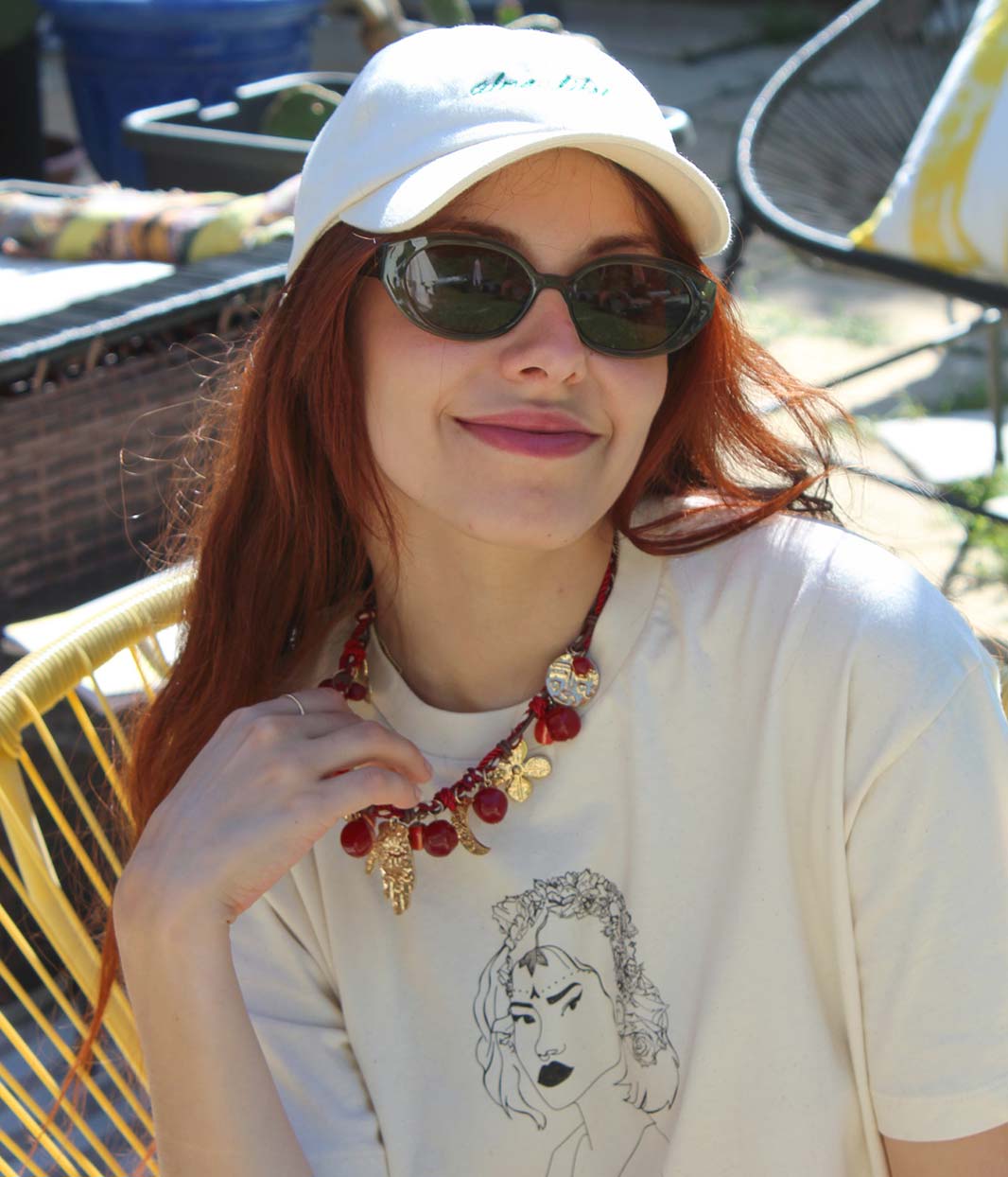 Femme rousse portant une casquette écrue et des lunettes noires, assise en extérieur dans un fauteuil jaune, avec un t-shirt écru illustré d’un portrait féminin noir.
