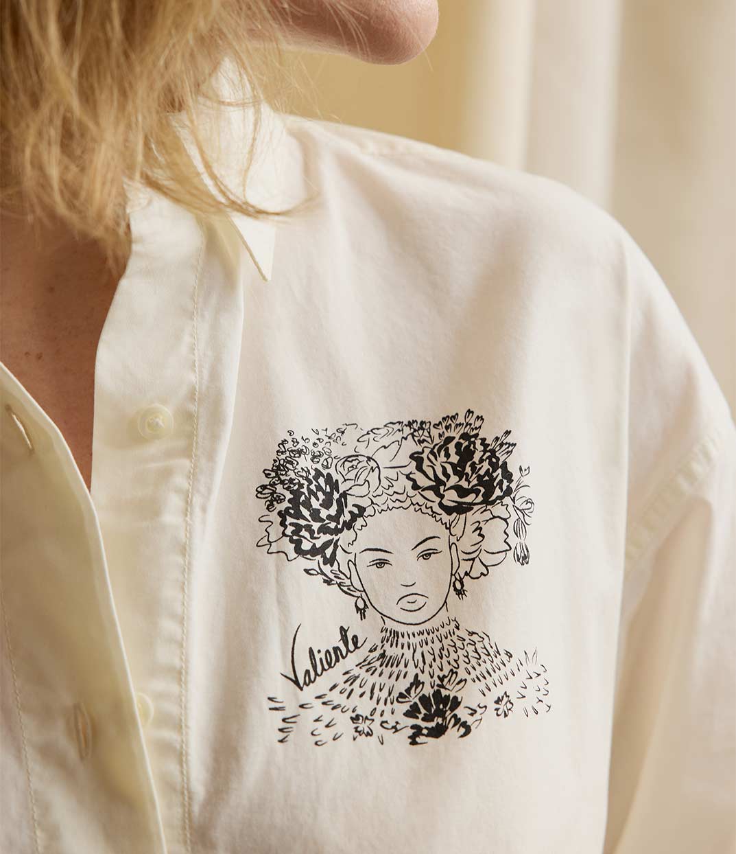 Gros plan sur une femme portant une chemise écru en coton biologique, imprimé noir d'une femme forte avec des fleurs dans les cheveux représentant la première reine nigériane accompagnée de l'inscription  "Valiente" sur le côté. Modèle Valiente conçu et imprimé à Lyon par la marque éthique Leonor Roversi.