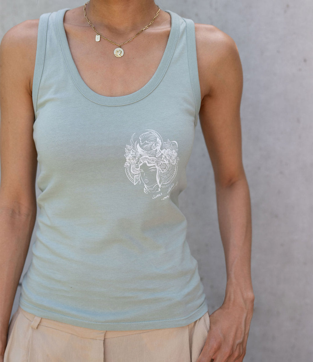 Vue rapprochée d’un débardeur pour femme en coton bio vert menthe. Il présente un imprimé blanc représentant une figure de samouraï couronnée de fleurs, accompagné de l’inscription « âme d’une conquérante ». Modèle Gozen, conçu et imprimé à Lyon par la marque éthique Leonor Roversi.