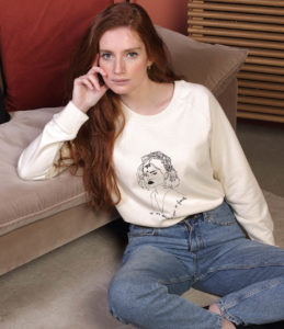 Une femme rousse porte un sweatshirt crème Lupita beige leonor roversi. Elle est assise et regarde l'objectif.