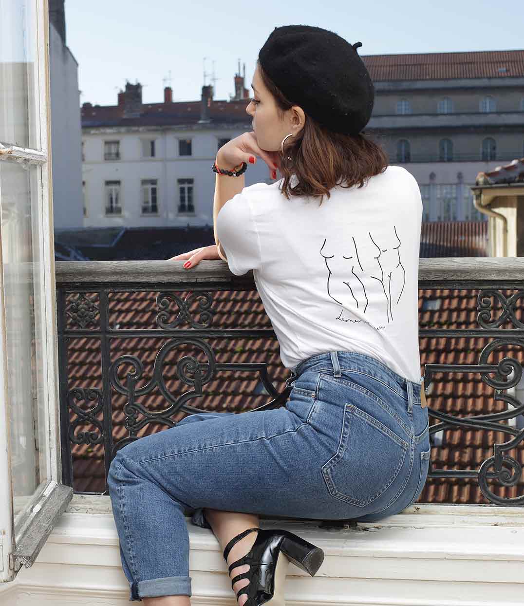 Une femme de dos et assis sur le bord d’une fenêtre portant un t-shirt Leonor Roversi marque lyonnaise féministe éthique et éco-responsable